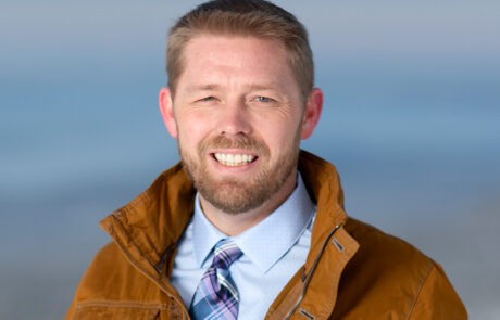 Josh Revak headshot, smiling in tie and brown jacket outside with a winter blue backdrop